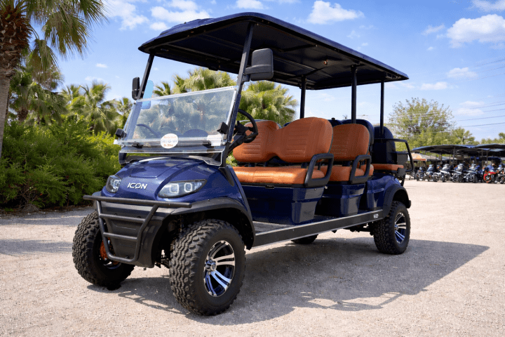Blue golf cart with orange seats parked on gravel with palm trees in the background.